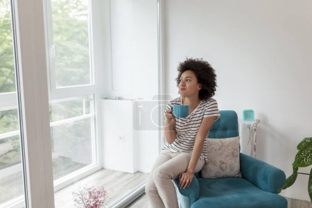 Foto de Hermosa mujer de raza mixta sentada en un sillón junto a la ventana, bebiendo café y disfrutando del tiempo libre en casa - Imagen libre de derechos