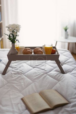 Téléchargez les photos : Plateau de petit déjeuner avec vase de fleurs, jus d'orange, croissants et café placés sur le lit. Focus sur les croissants - en image libre de droit