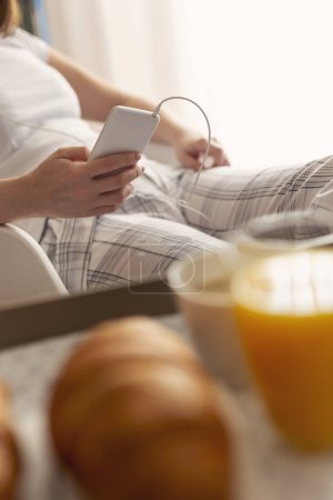 Photo for Detail of a woman wearing pajamas, sitting in bedroom, holding a mobile phone and listening to the music. Breakfast tray in the foreground. Focus on the phone - Royalty Free Image
