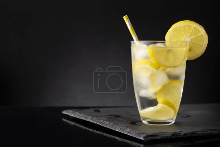 Foto de Un vaso de agua fresca y fría con rodajas de limón, cubitos de hielo y una pajita para beber en una bandeja de piedra negra - Imagen libre de derechos