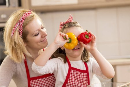 Foto de Hermosa madre e hija divirtiéndose haciendo ensalada en la cocina, sosteniendo rodajas de pimienta como vasos y mirando. Concéntrate en la hija - Imagen libre de derechos