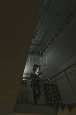 Téléchargez les photos : Belle jeune femme debout sur un escalier dans un couloir de bâtiment à faible luminosité, tapant un message texte à l'aide d'un téléphone intelligent - en image libre de droit