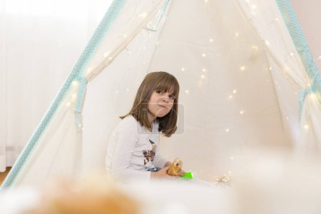 Foto de Niña jugando en una tienda de campaña en su sala de juegos, sosteniendo una linterna y comiendo croissant para el desayuno - Imagen libre de derechos