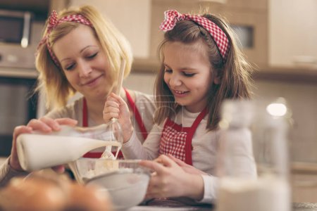 Photo for Beautiful mother and daughter having fun in the kitchen; mother pouring milk while daughter is kneading dough, stiring flour with kitchen spoon. Focus on the daughter - Royalty Free Image