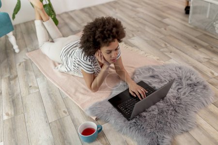 Téléchargez les photos : Vue en angle élevé d'une pigiste couchée sur le sol du salon, travaillant sur un ordinateur portable et buvant du café ; femme travaillant à distance depuis la maison - en image libre de droit