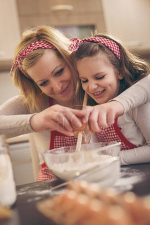 Téléchargez les photos : Jeune mère dans la cuisine cuisson pâte avec sa fille, casser un oeuf tandis que la fille est en remuant la farine - en image libre de droit