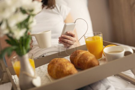 Foto de Detalle de una mujer con pijama, sentada en la cama, tomando café y escuchando música. Bandeja de desayuno bellamente servido en primer plano. Concéntrate en la parte superior del teléfono - Imagen libre de derechos