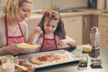 Photo for Mother and daughter in the kitchen making pizza, putting the cheese on the pizza dough. Focus on the daughter - Royalty Free Image