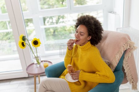 Photo for Beautiful young mixed race woman relaxing at home, sitting in an armchair and eating a fresh croissant for breakfast - Royalty Free Image