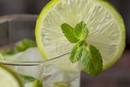 Foto de Detalle de vaso de cóctel mojito con mucho hielo, ron blanco, zumo de limón y tónico, decorado con rodajas de lima y hojas de menta - Imagen libre de derechos