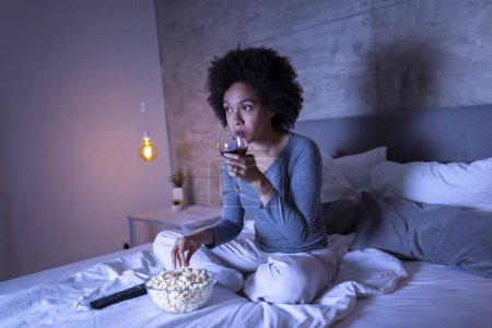 Téléchargez les photos : Belle femme métisse assise sur le lit, buvant du vin, mangeant du pop-corn et regardant la télévision - en image libre de droit