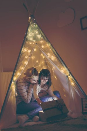 Foto de Madre e hija sentadas en un tipi iluminadas por las luces de Navidad, abriendo un regalo. Concéntrate en la madre - Imagen libre de derechos