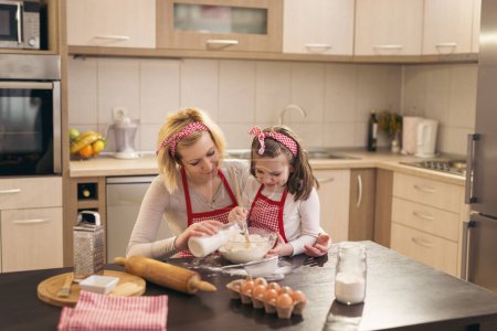 Téléchargez les photos : Belle mère et fille s'amusent dans la cuisine ; mère versant du lait pendant que la fille pétrit la pâte, en remuant la farine avec une cuillère de cuisine. Focus sur la mère - en image libre de droit