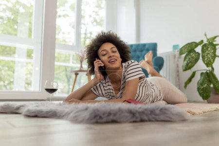 Photo for Portrait of a beautiful young woman lying on the floor, having a phone conversation, drinking wine and relaxing at home - Royalty Free Image