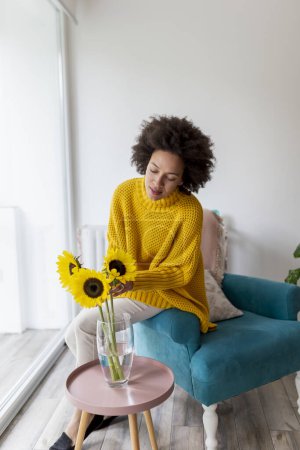 Photo for Beautiful young mixed race woman relaxing at home, putting sunflower bouquet into a vase - Royalty Free Image