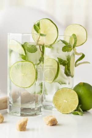 Téléchargez les photos : Deux cocktails mojito avec beaucoup de glace, rhum blanc, jus de citron et tonique, décorés de tranches de citron vert et de feuilles de menthe sur une table blanche moderne. - en image libre de droit