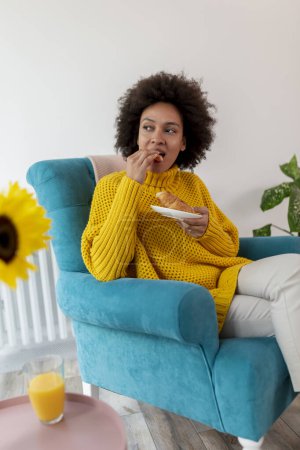 Foto de Hermosa mujer de raza mixta relajándose en casa, sentada en un sillón y desayunando - Imagen libre de derechos