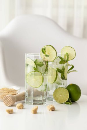 Téléchargez les photos : Deux cocktails mojito avec beaucoup de glace, rhum blanc, jus de citron et tonique, décorés de tranches de citron vert et de feuilles de menthe sur une table blanche moderne - en image libre de droit