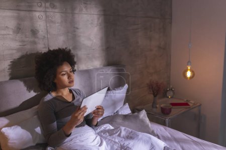 Téléchargez les photos : Belle jeune femme de race mixte portant un pyjama couché dans le lit le matin, lisant un livre électronique à l'aide d'un ordinateur tablette et se détendre à la maison - en image libre de droit