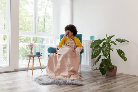 Foto de Retrato de una mujer enferma y cansada sentada en un sillón cubierto de manta, con migraña, sosteniendo la cabeza en las manos - Imagen libre de derechos