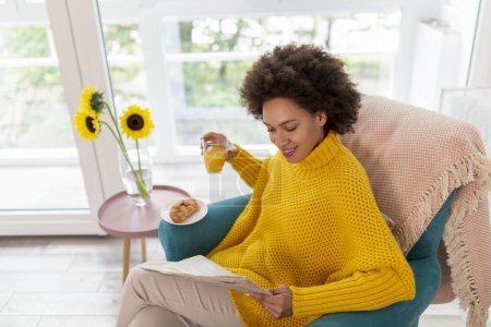 Foto de Hermosa mujer de raza mixta relajándose en casa, sentada en un sillón, leyendo periódicos y bebiendo jugo de naranja - Imagen libre de derechos
