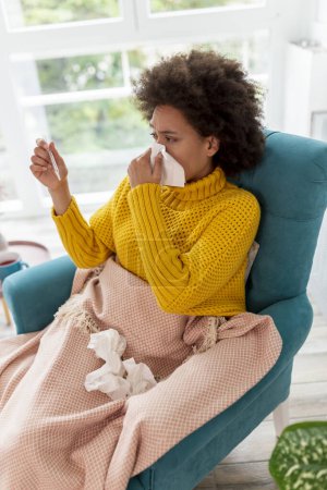 Téléchargez les photos : Portrait de femme malade assise dans un fauteuil recouvert d'une couverture, ayant une grippe et de la fièvre, se mouchant dans un mouchoir en papier - en image libre de droit
