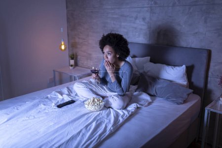 Foto de Hermosa mujer de raza mixta sentada en la cama, bebiendo vino, comiendo palomitas de maíz y viendo una película en la televisión - Imagen libre de derechos