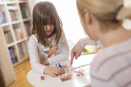 Photo for Mother and daughter sitting in a playroom, playing a ludo game; daughter repositioning the pawn - Royalty Free Image