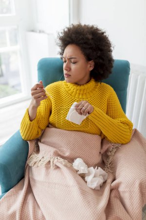Foto de Retrato de una mujer enferma sentada en un sillón cubierto con manta, con gripe y fiebre, estornudos y sonando la nariz en un pañuelo de papel - Imagen libre de derechos