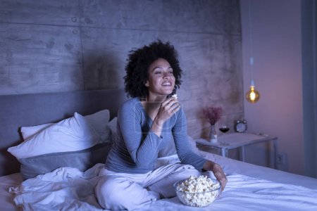 Téléchargez les photos : Belle femme de race mixte portant un pyjama assis sur le lit, mangeant du pop-corn et regardant un film comique à la télévision, se détendre à la maison tard dans la nuit - en image libre de droit