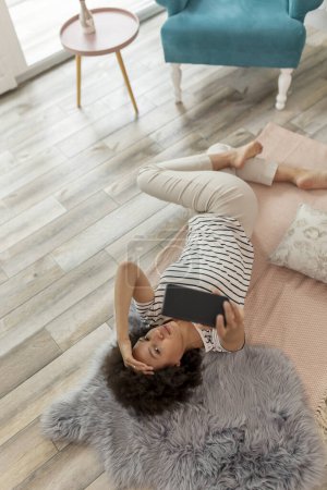 Foto de Vista superior de una hermosa joven mestiza tumbada en el suelo de la sala de estar, tomando un slefie con su teléfono inteligente y divirtiéndose mientras se relaja en casa - Imagen libre de derechos