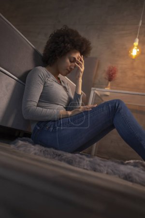 Téléchargez les photos : Belle jeune femme métisse assise par terre près du lit les yeux fermés, triste et stressée - en image libre de droit