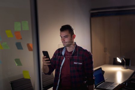 Téléchargez les photos : Développeur logiciel travaillant tard dans un bureau, debout dans une salle de conférence à côté d'un mur de verre avec des notes collantes pour le remue-méninges et l'utilisation d'un smartphone - en image libre de droit