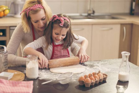 Téléchargez les photos : Biscuits à la mère et à la fille dans la cuisine ; petite fille utilisant un rouleau à pâtisserie, pétrissant la pâte à biscuits - en image libre de droit