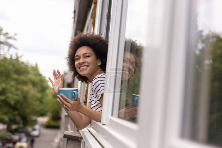 Téléchargez les photos : Portrait d'une belle femme métissée debout près de la fenêtre et regardant dans la rue, saluant un ami, se relaxant à la maison et buvant du café - en image libre de droit