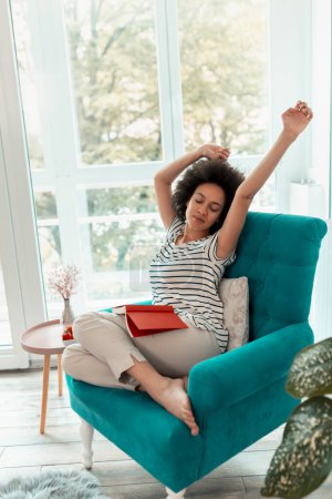Photo for Beautiful young mixed race woman stretching while sitting in an armchair by the window, reading a book and enjoying leisure time at home - Royalty Free Image