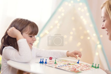 Photo for Mother and daughter sitting in a playroom, playing a ludo game; daughter repositioning the pawn - Royalty Free Image