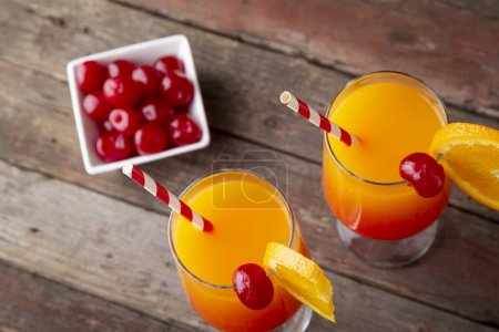 Photo for High angle view of two cold tequila sunrise cocktails with tequila, pomegranate juice and orange juice decorated with slices of orange and maraschino cherries. Focus on the tips of the straw - Royalty Free Image