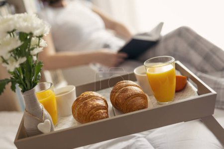 Foto de Mujer con pijama, sentada en una silla al lado de la cama, leyendo un libro. Bandeja de desayuno en primer plano. Enfoque selectivo en el croissant derecho - Imagen libre de derechos