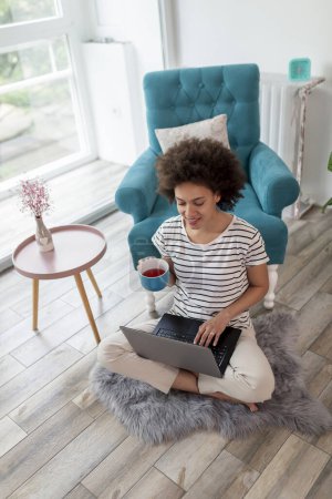 Foto de Hermosa joven mujer de raza mixta sentado en el piso de la sala de estar, tener una videollamada en una computadora portátil y beber té - Imagen libre de derechos