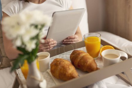 Foto de Detalle de las manos de la hembra sosteniendo una tableta negra con pantalla blanca en blanco, sentada en la cama y desayunando. Enfoque selectivo en la mano izquierda - Imagen libre de derechos