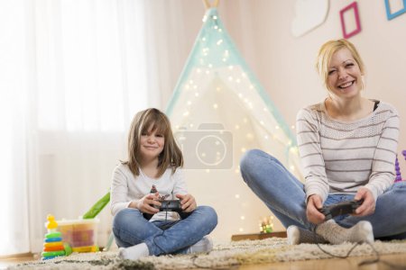 Téléchargez les photos : Mère et fille assises sur le sol dans une salle de jeux, jouant à des jeux vidéo et s'amusant. Focus sur la fille - en image libre de droit