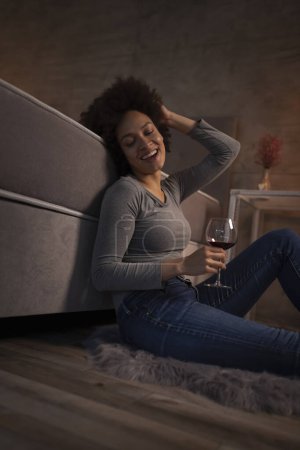Foto de Hermosa joven mujer de raza mixta sentada en el suelo junto a la cama, tomando una copa de vino y relajándose en casa después de un día ajetreado - Imagen libre de derechos