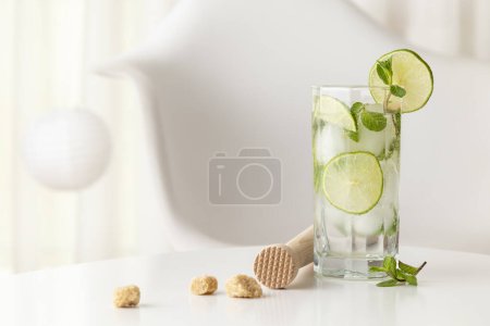 Téléchargez les photos : Cocktail mojito avec beaucoup de glace, rhum blanc, jus de citron et tonique, décoré de tranches de citron vert et de feuilles de menthe sur une table blanche moderne - en image libre de droit