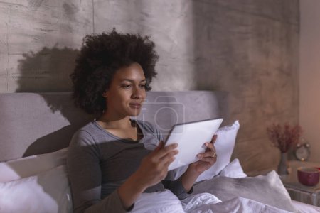 Téléchargez les photos : Belle jeune femme métisse en pyjama, couchée au lit le matin, vérifiant les e-mails à l'aide d'un ordinateur tablette et se relaxant à la maison - en image libre de droit