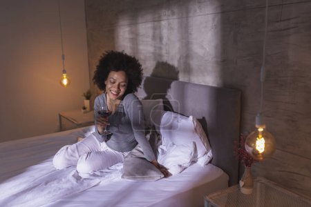 Foto de Mujer con pijama sentada en la cama, bebiendo vino y disfrutando del tiempo libre en casa - Imagen libre de derechos