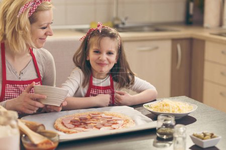 Foto de Madre e hija en la cocina haciendo pizza, poniendo el salami en la masa de pizza. Concéntrate en la hija - Imagen libre de derechos