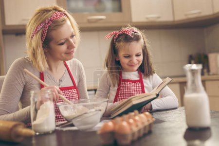 Téléchargez les photos : Mère et fille font des biscuits dans la cuisine, à la recherche d'une recette dans un livre de cuisine. Focus sur la fille - en image libre de droit