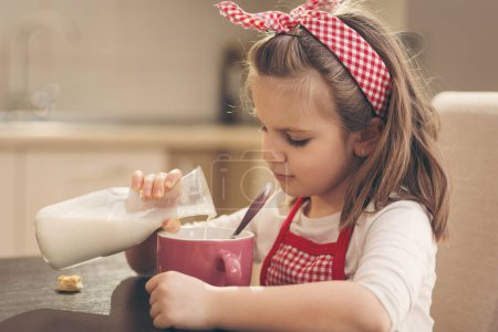 Téléchargez les photos : Petite fille assise dans la cuisine, prenant le petit déjeuner, versant du lait dans une tasse de céréales, se préparant un repas sain et savoureux - en image libre de droit