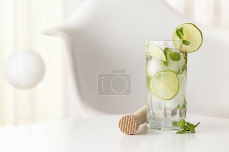 Foto de Cóctel Mojito con mucho hielo, ron blanco, zumo de limón y tónico, decorado con rodajas de lima y hojas de menta en una moderna mesa blanca - Imagen libre de derechos